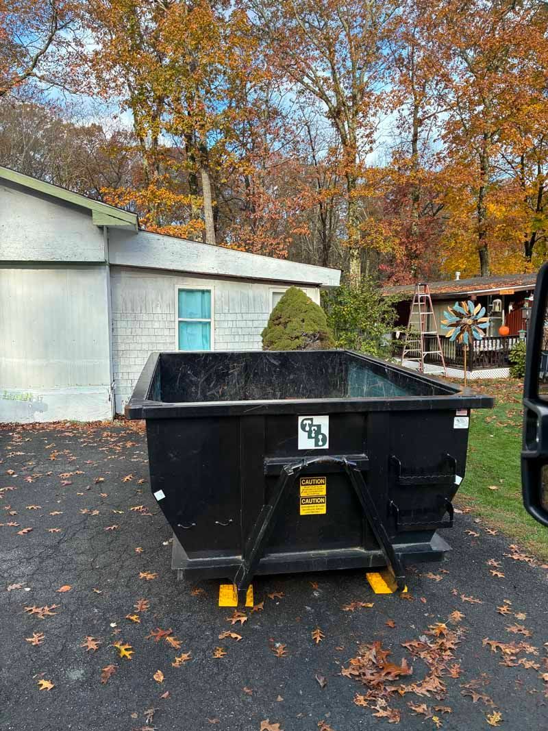 Black dumpster on a wet driveway beside a white building, with autumn trees in the background.