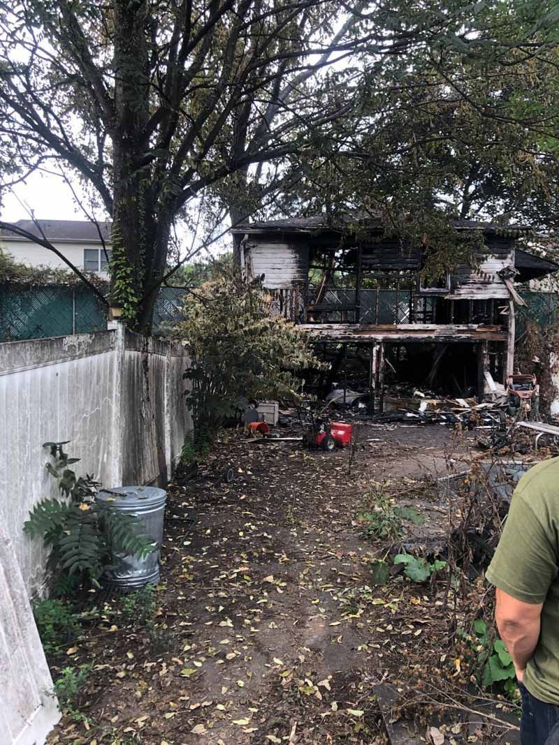 Backyard with trees, concrete wall, and a partially burned shed or house at the rear.