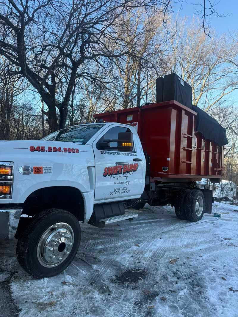 White dump truck with a red bed parked on a snowy road beside bare trees