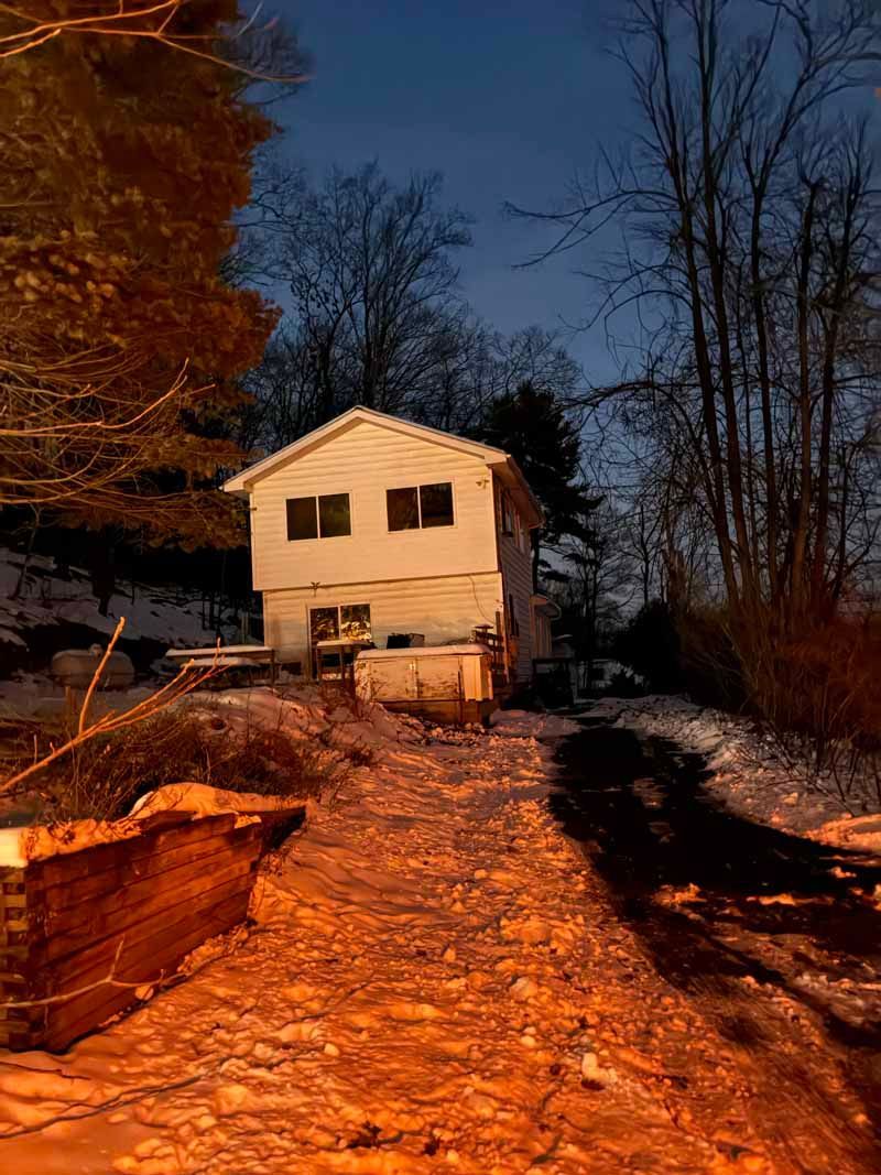 Snowy dirt path leading to a small white house at dusk, lit by warm orange light, with bare trees nearby