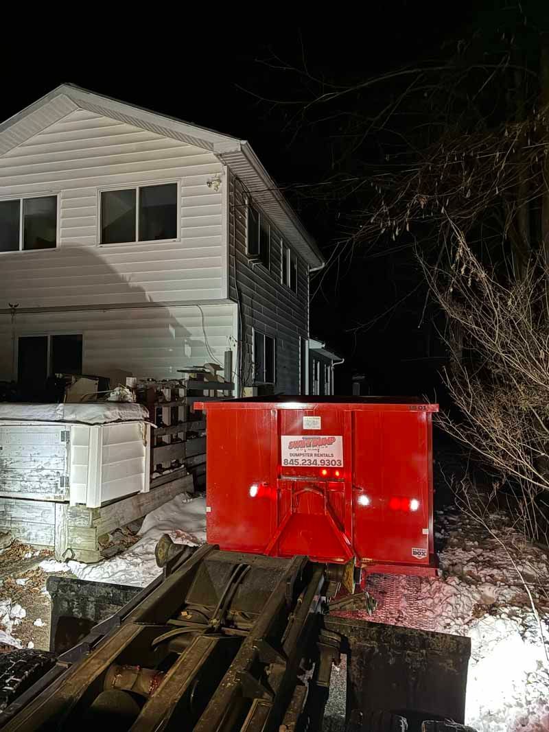 Red dumpster beside a house at night, with wooden planks in the foreground and snow on the ground.