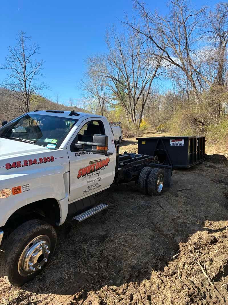 White dump truck with trailer parked on a dirt path beside a wooded area under a clear blue sky