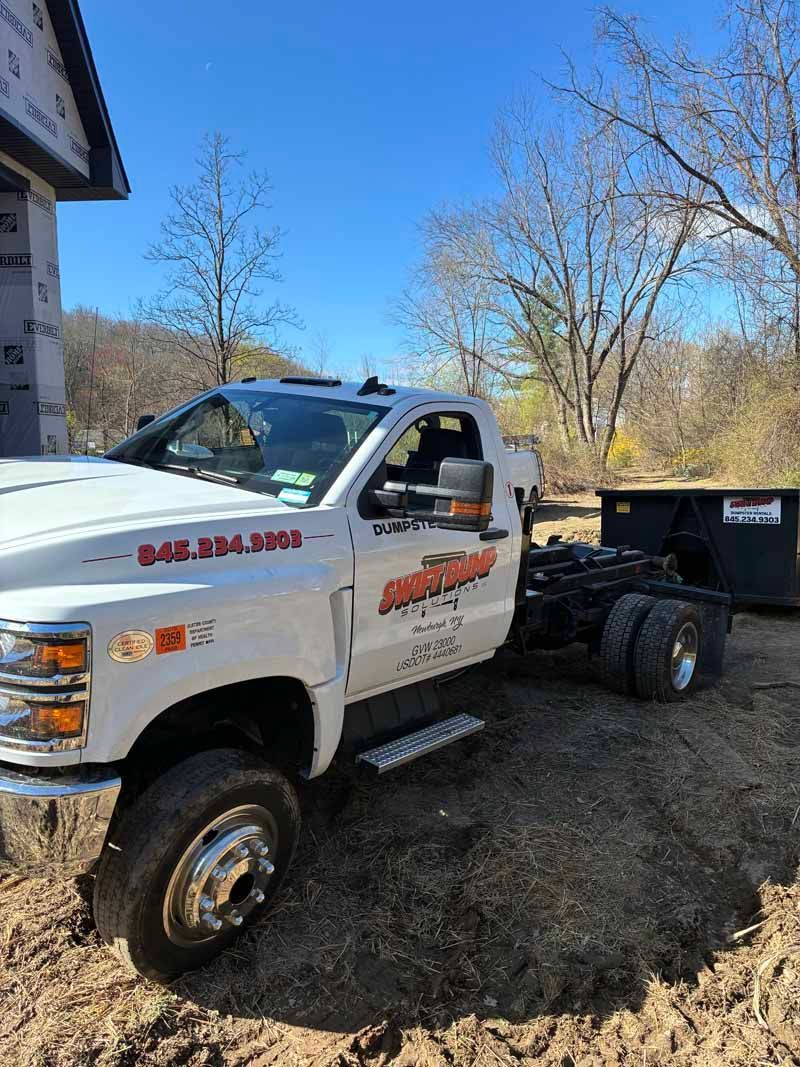 White dump truck with a black trailer parked beside a house on a sunny dirt lot