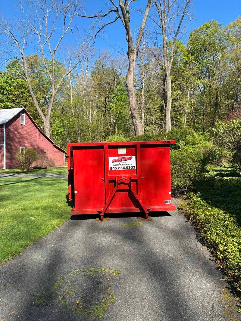 Red dumpster blocking a driveway beside a red barn in a grassy yard with trees