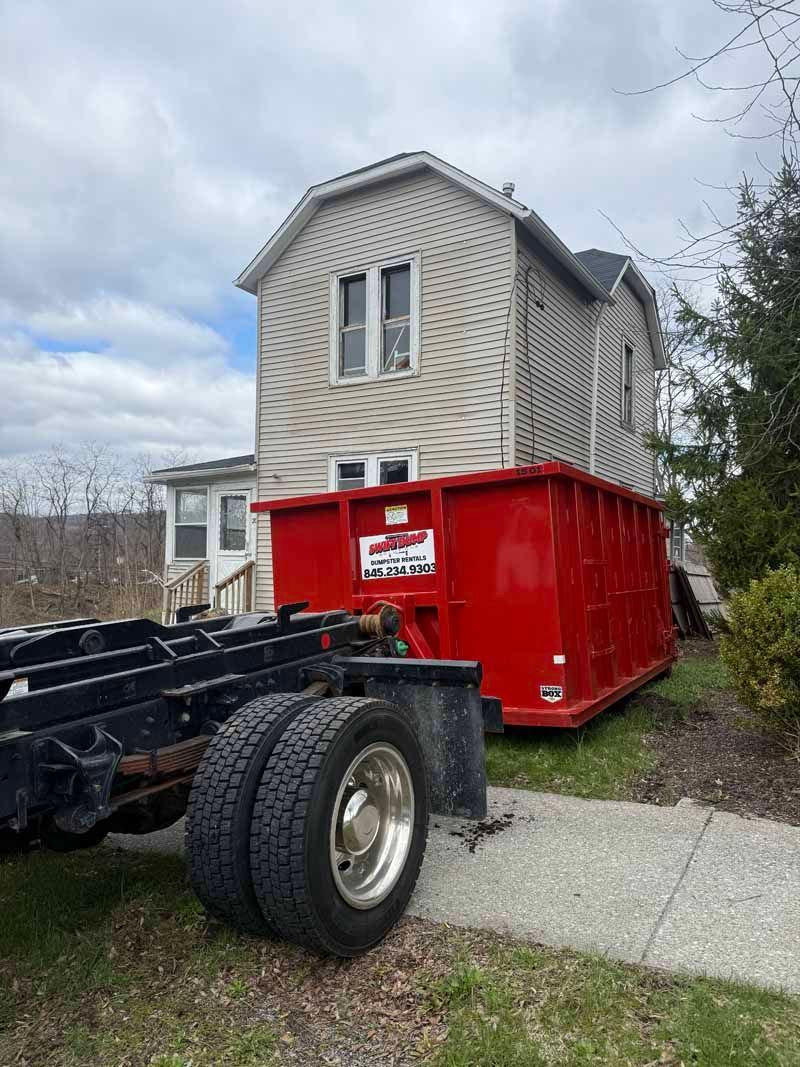 Truck chassis with a red dumpster beside a gray house on a gravel driveway