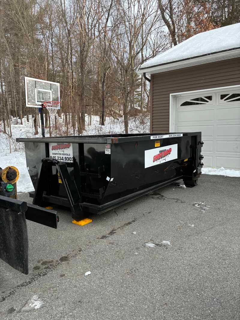 Black dumpster on gravel driveway beside a garage and outdoor basketball hoop, with snow on the roof.