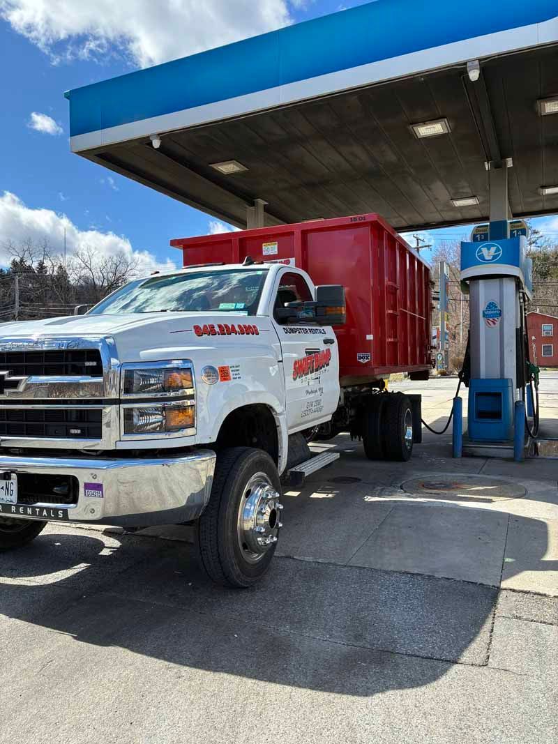 White semi truck loading red dumpster at a blue-canopy fuel station