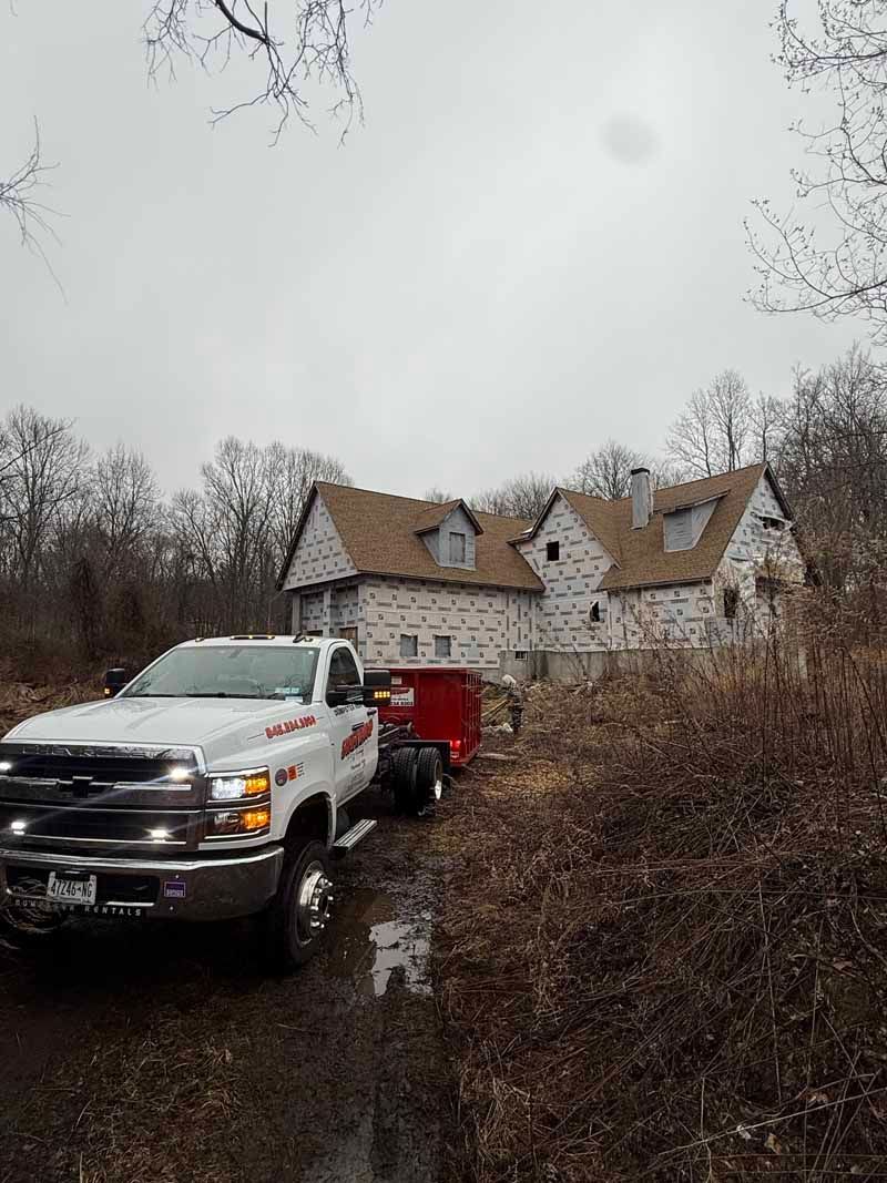 White pickup truck with red trailer on muddy path beside an unfinished house in a wooded area