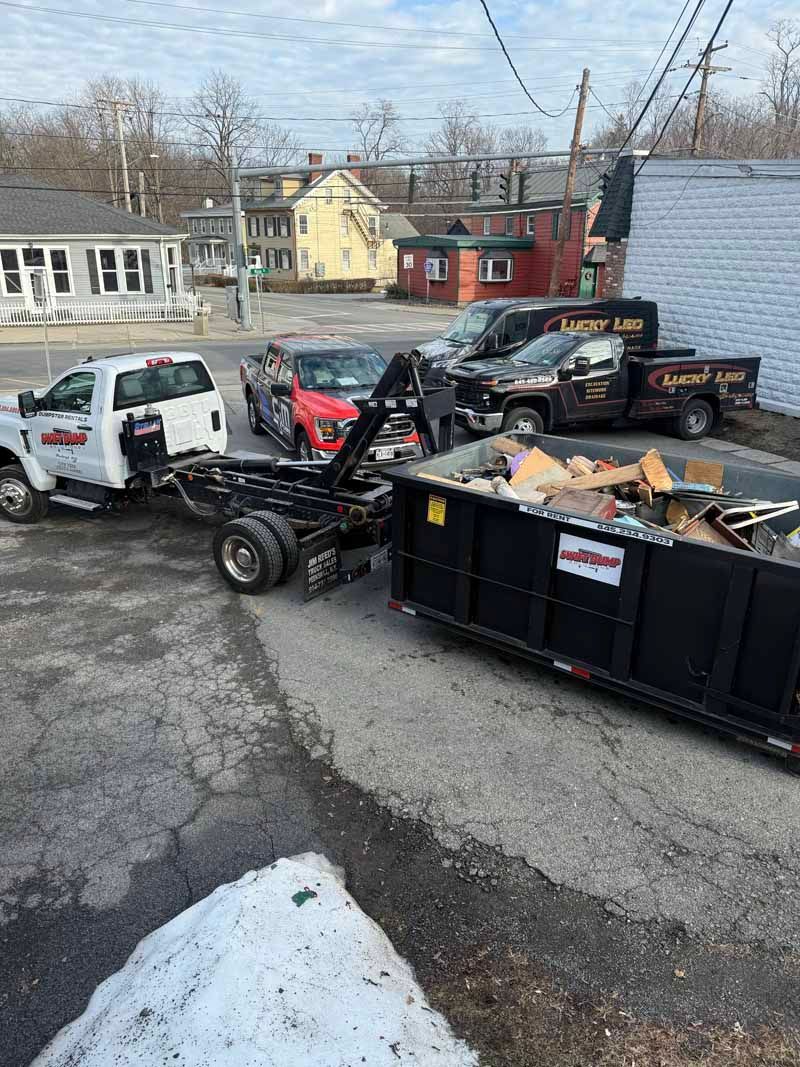 Two tow trucks at a roadside dump site with a black dumpster and scattered debris in winter