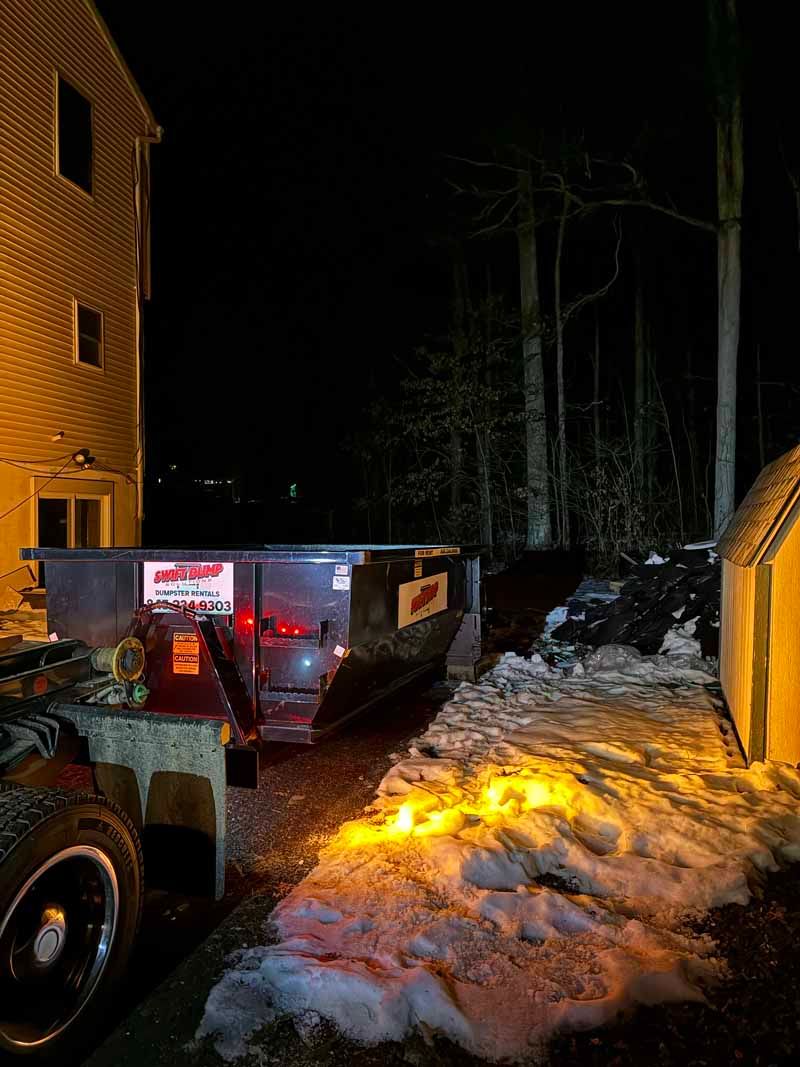 Snow-covered driveway with a tow truck and trailer beside a brick building at night