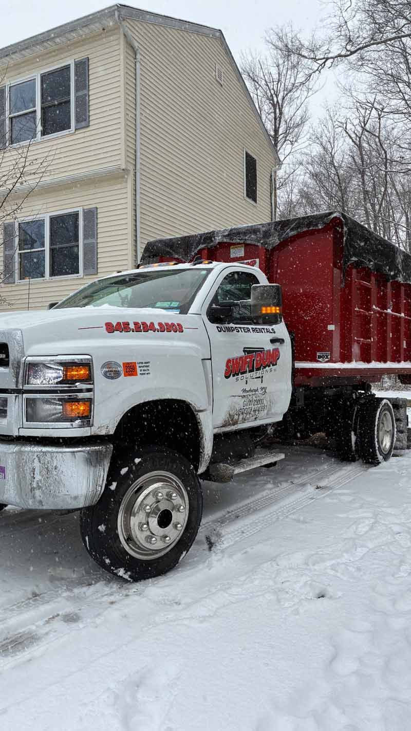 White tow truck with red dump bed in snowy street beside a beige house in winter