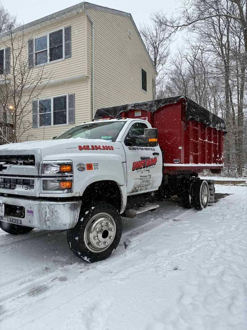 White dump truck hauling red trailer on a snowy driveway beside a beige house