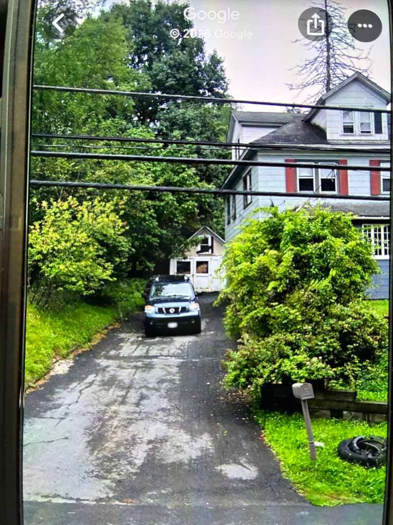 Narrow driveway with a parked silver car beside a red house and lush green trees.