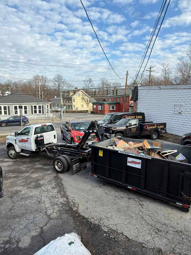 Construction debris in dumpsters beside parked work trucks in a roadside lot under a blue sky