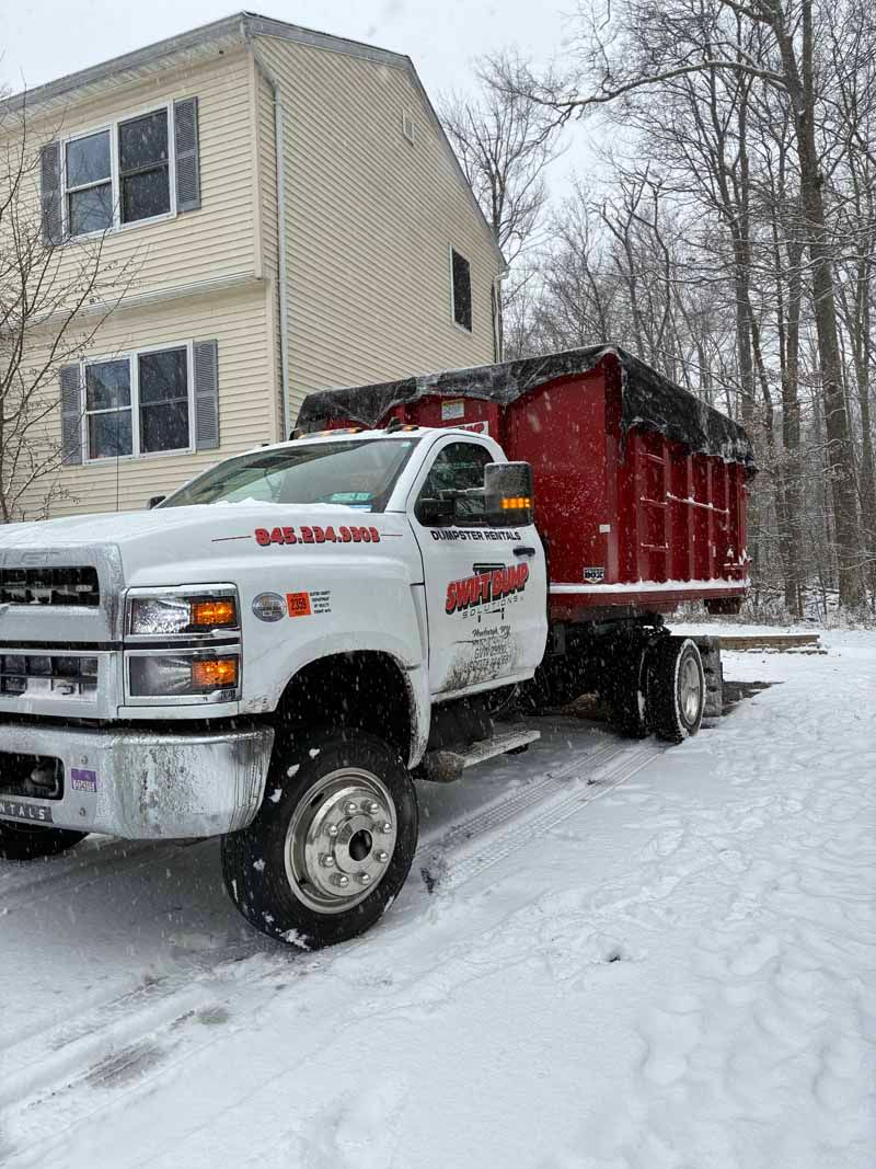 White dump truck with red trailer parked in snowy driveway beside a beige house.