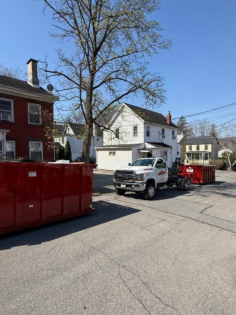 Street scene with white pickup truck, red dumpsters, and houses under a clear blue sky