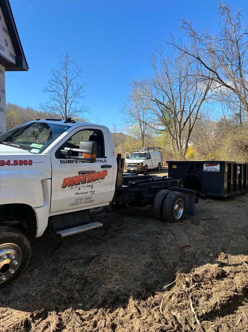 White work truck hauling a black dumpster in a muddy yard under a clear blue sky