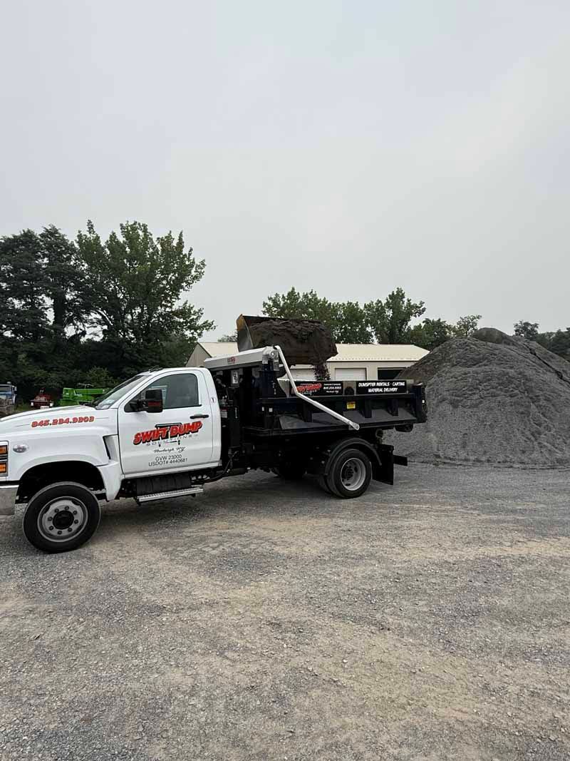 White dump truck at a gravel site, dumping material beside a large pile under an overcast sky