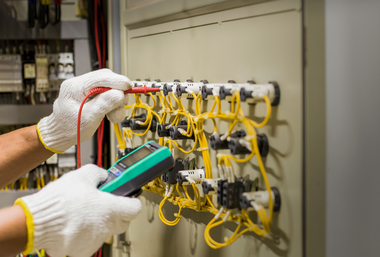 Gloved hands testing a circuit panel with yellow wires and a green multimeter probe
