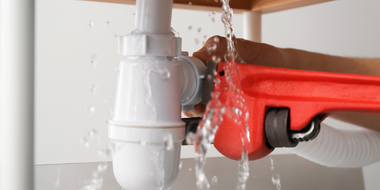 Red pipe clamp holding a white drain pipe while water leaks into a sink cabinet