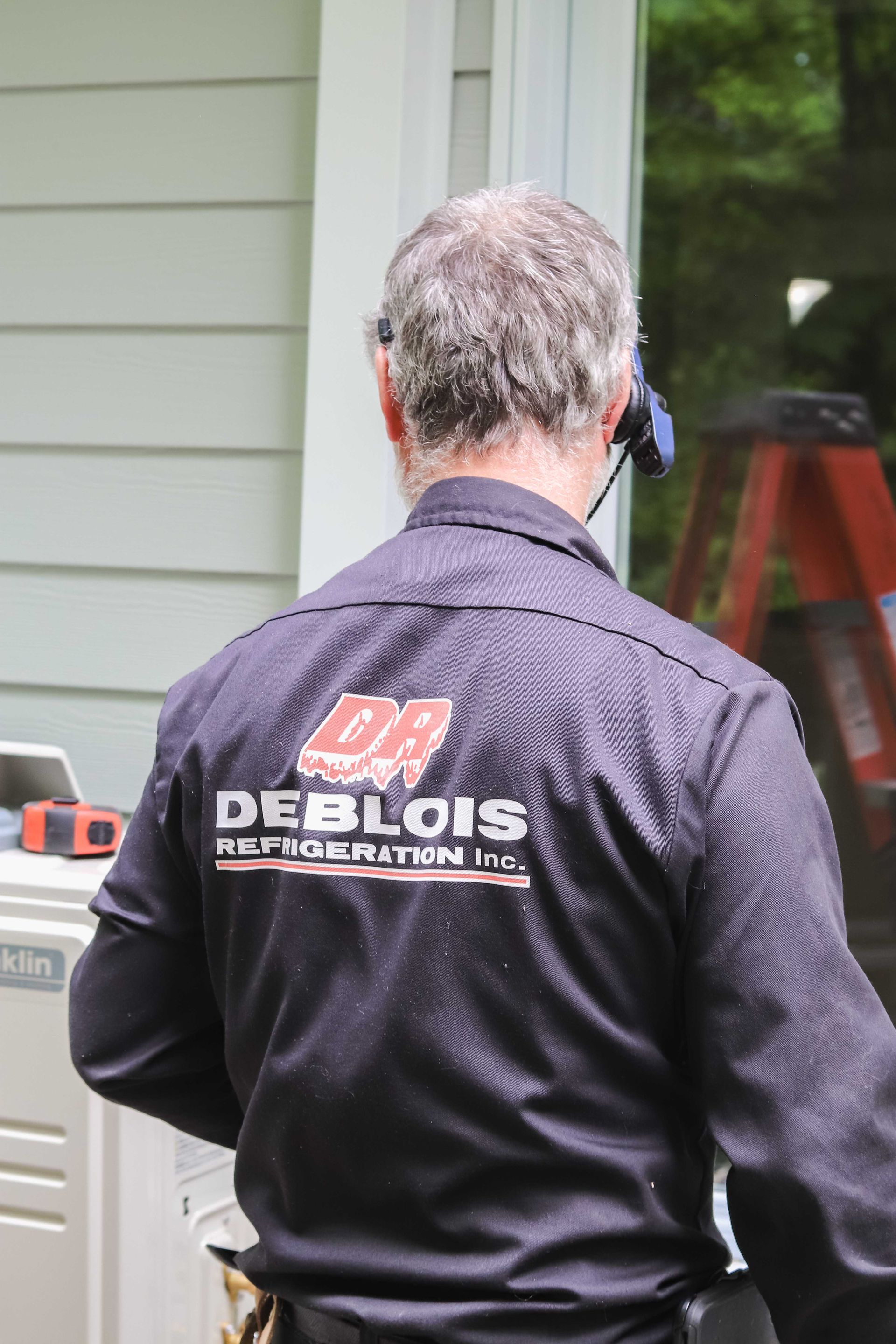 Un homme portant une chemise de travail bleu foncé avec le logo « Deblois Roof Inspection Inc. » se tient près d'un cadre de fenêtre et d'un revêtement blancs.