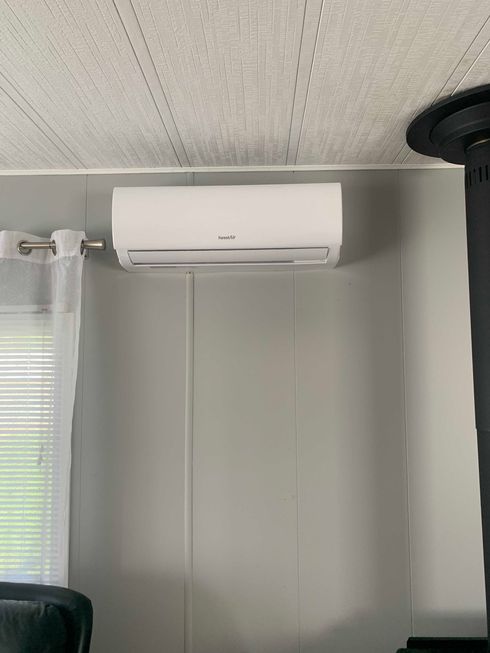 Climatiseur mural blanc sur un mur gris. Une fenêtre avec stores et rideaux se trouve à gauche.