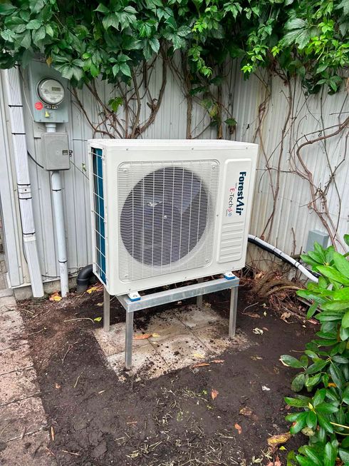 Unité de climatisation sur un support métallique, contre une clôture végétalisée.
