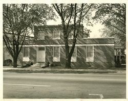 Brick building with two floors, flanked by trees, near a road.