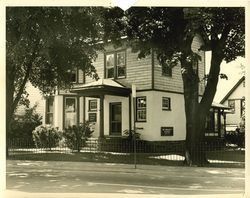 Black and white photo of a two-story house with a porch, trees in the front yard, and a fence along the street.
