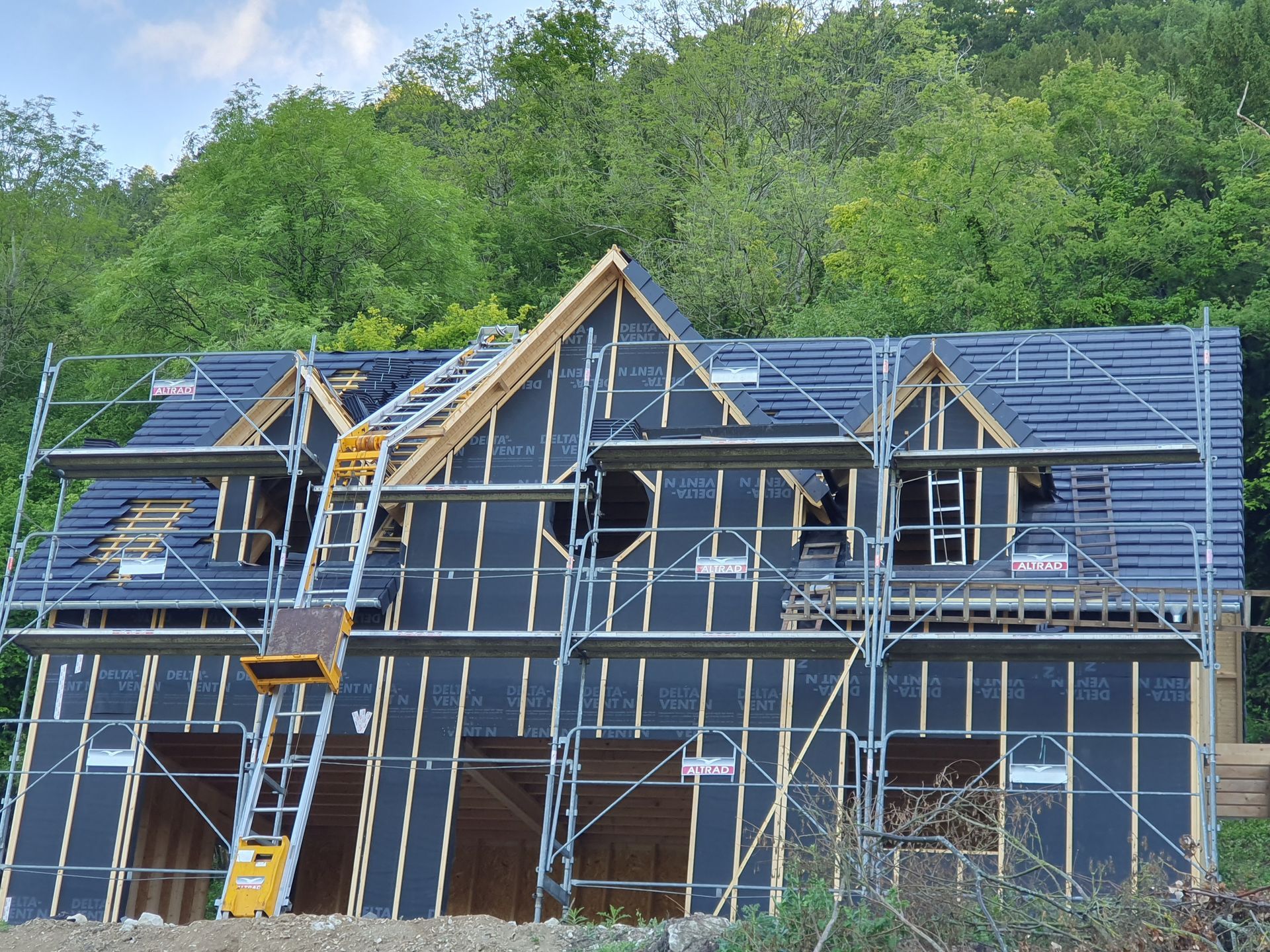Construction d'une maison à deux étages avec échafaudages, toit partiellement recouvert de bardeaux, sur fond d'arbres.