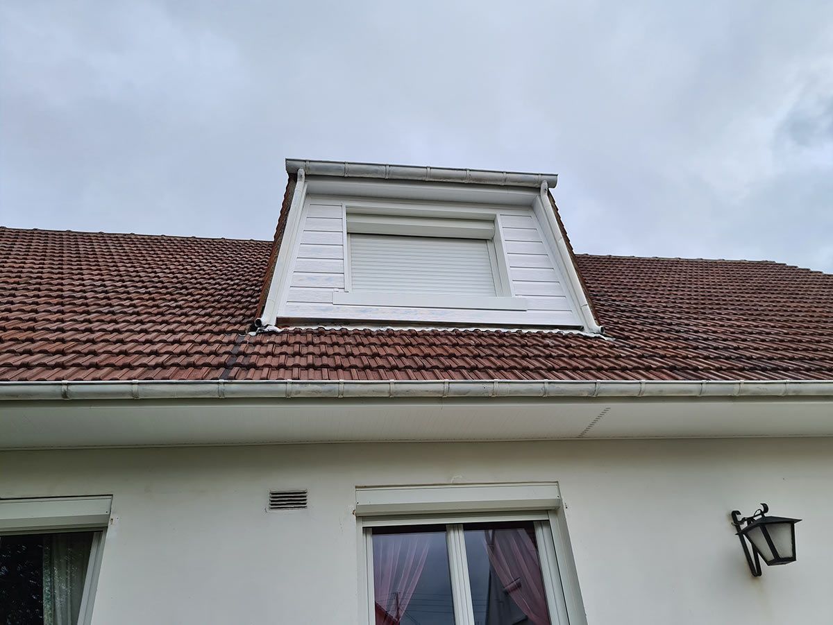 Maison blanche avec lucarne, toit de tuiles brunes et ciel couvert.