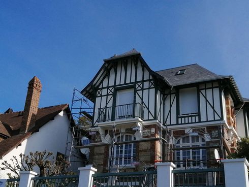 Maison à deux étages avec une charpente en bois noir et blanc, des accents de briques rouges et un ciel bleu en arrière-plan.