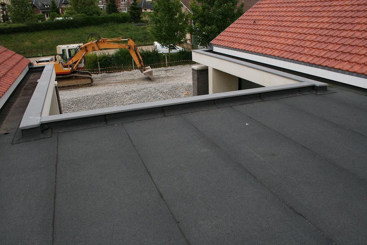 Toiture plate en bitume gris foncé jouxtant un toit de tuiles, avec un chantier et une excavatrice en arrière-plan.
