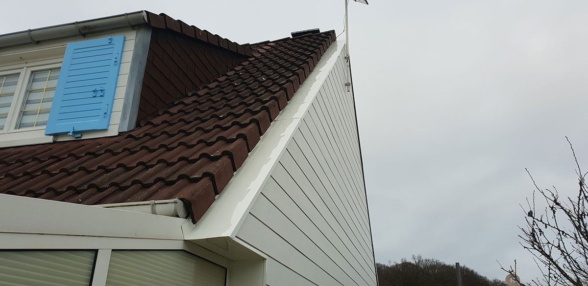 Une maison au toit de tuiles brunes et aux volets bleus se détachant sur un ciel nuageux.