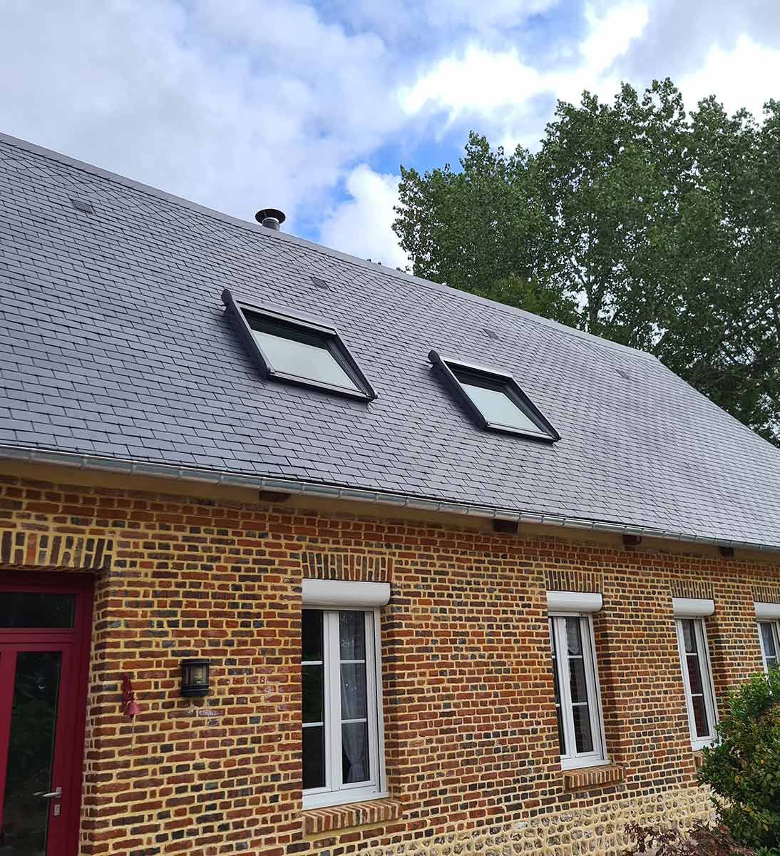 Maison en briques avec toit en ardoise grise, deux puits de lumière, fenêtres à cadre blanc et porte rouge.
