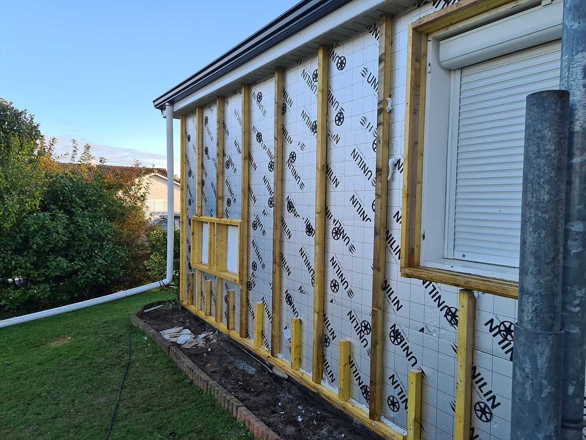 Rénovation d'un mur extérieur montrant des tasseaux de bois recouvrant une feuille d'isolation fixée à une maison