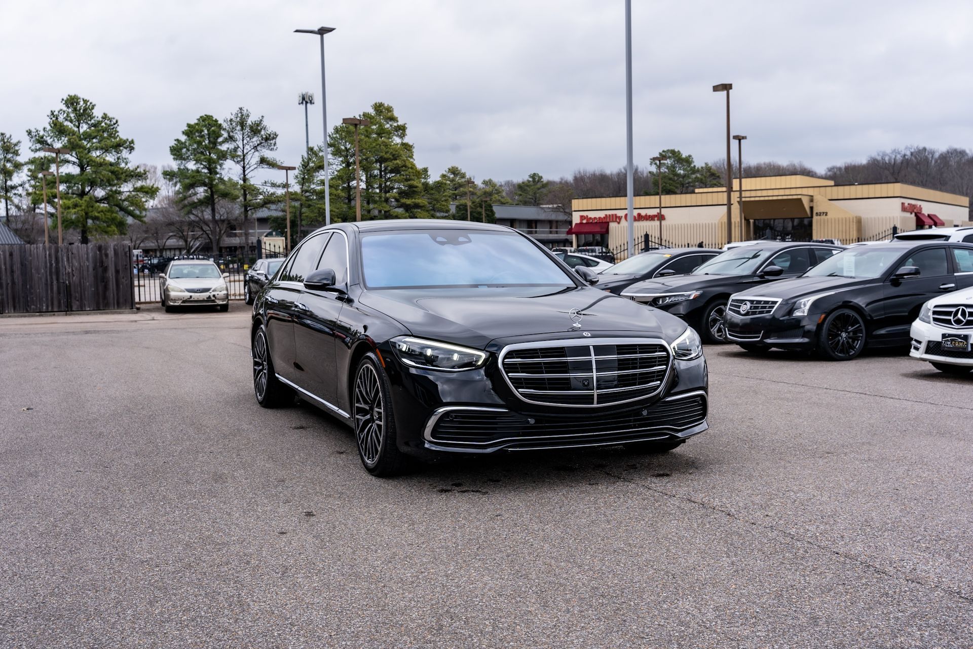 Black Mercedes-Benz sedan parked in a lot, surrounded by other cars under a cloudy sky.