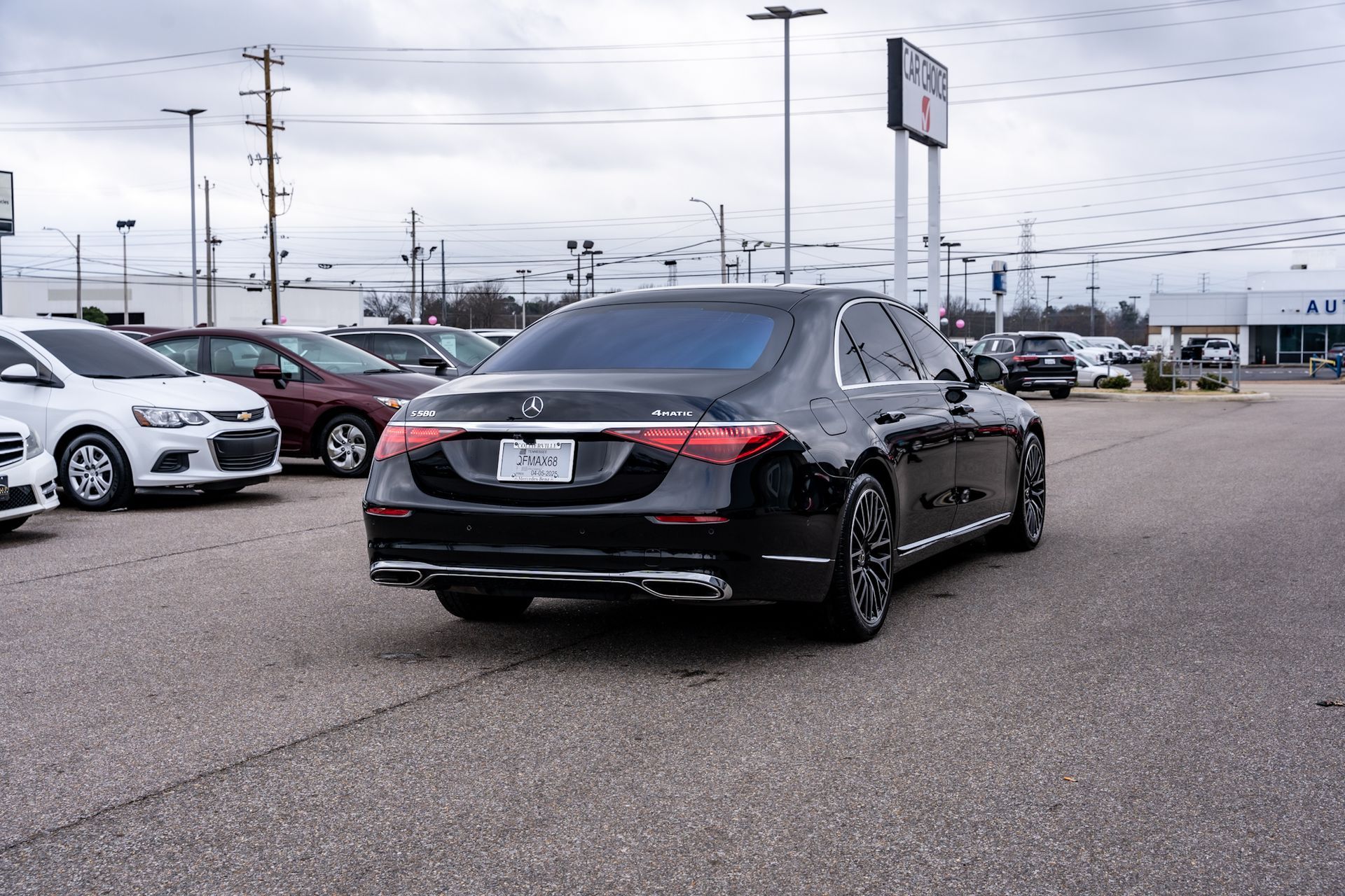 Black Mercedes-Benz sedan in a car dealership lot with other vehicles, under cloudy skies.