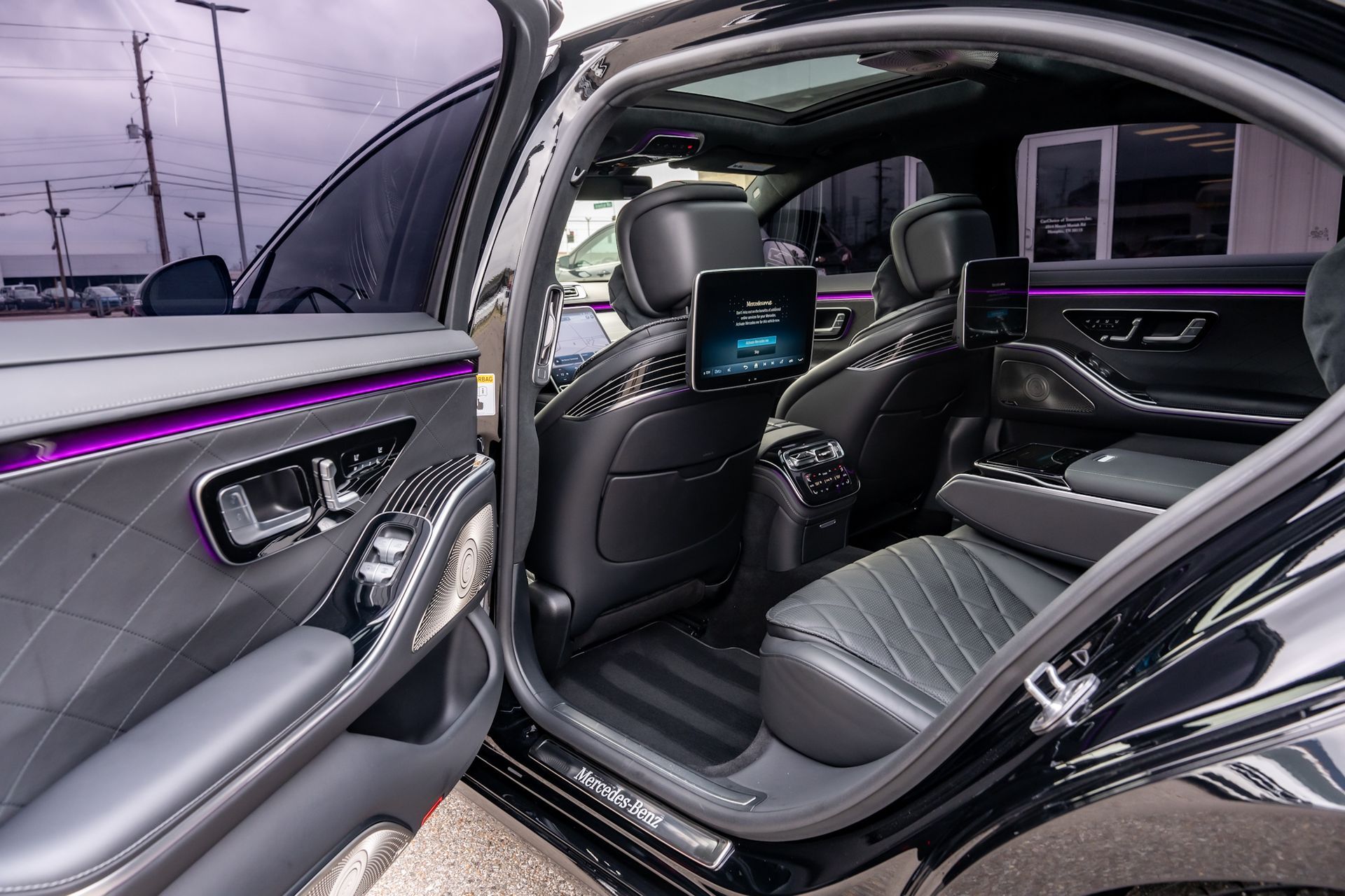 Interior of a black luxury car with open door, showing rear seating and screens; purple ambient lighting.
