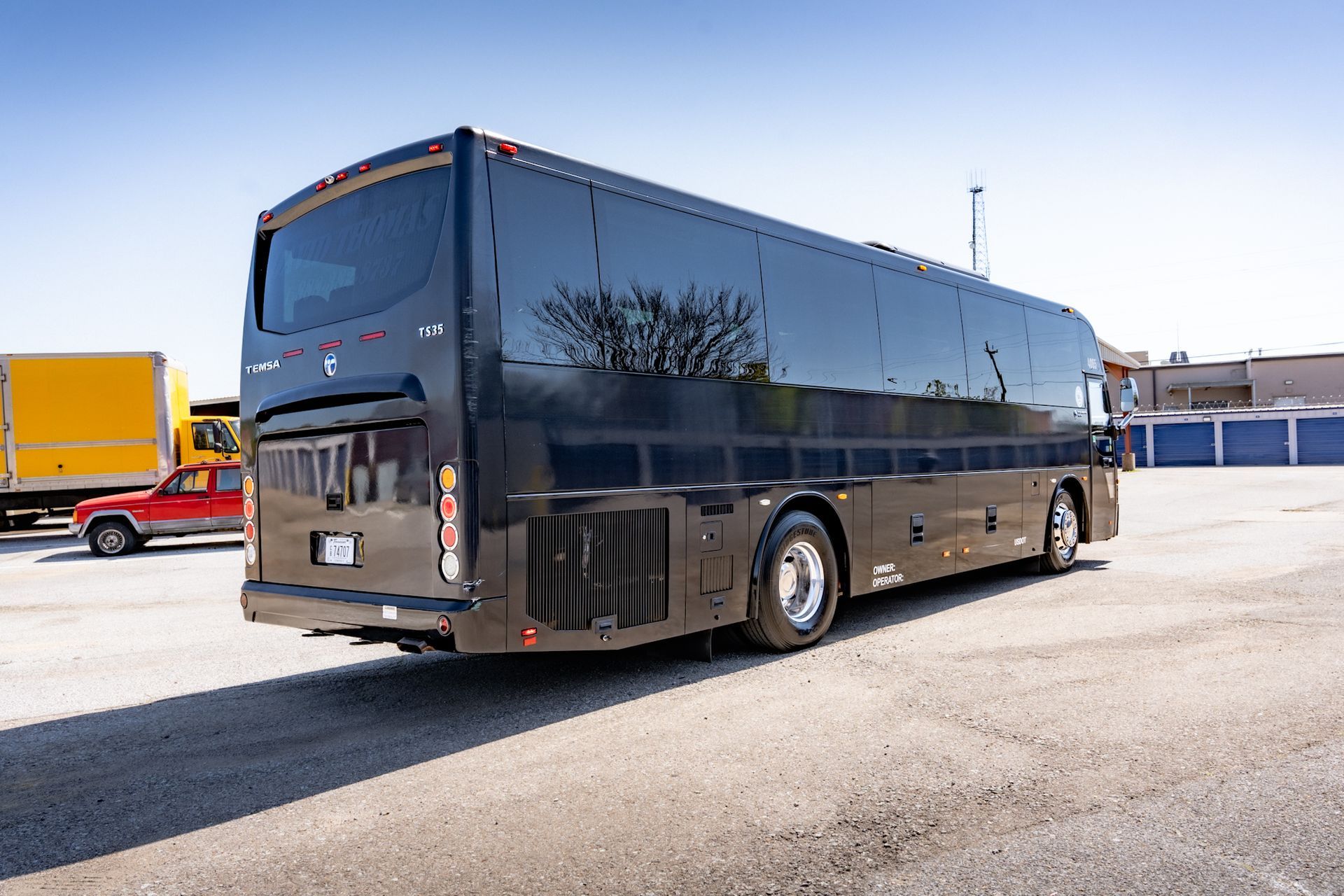 Black passenger bus parked on an asphalt lot; parked vehicles and storage units in the background.