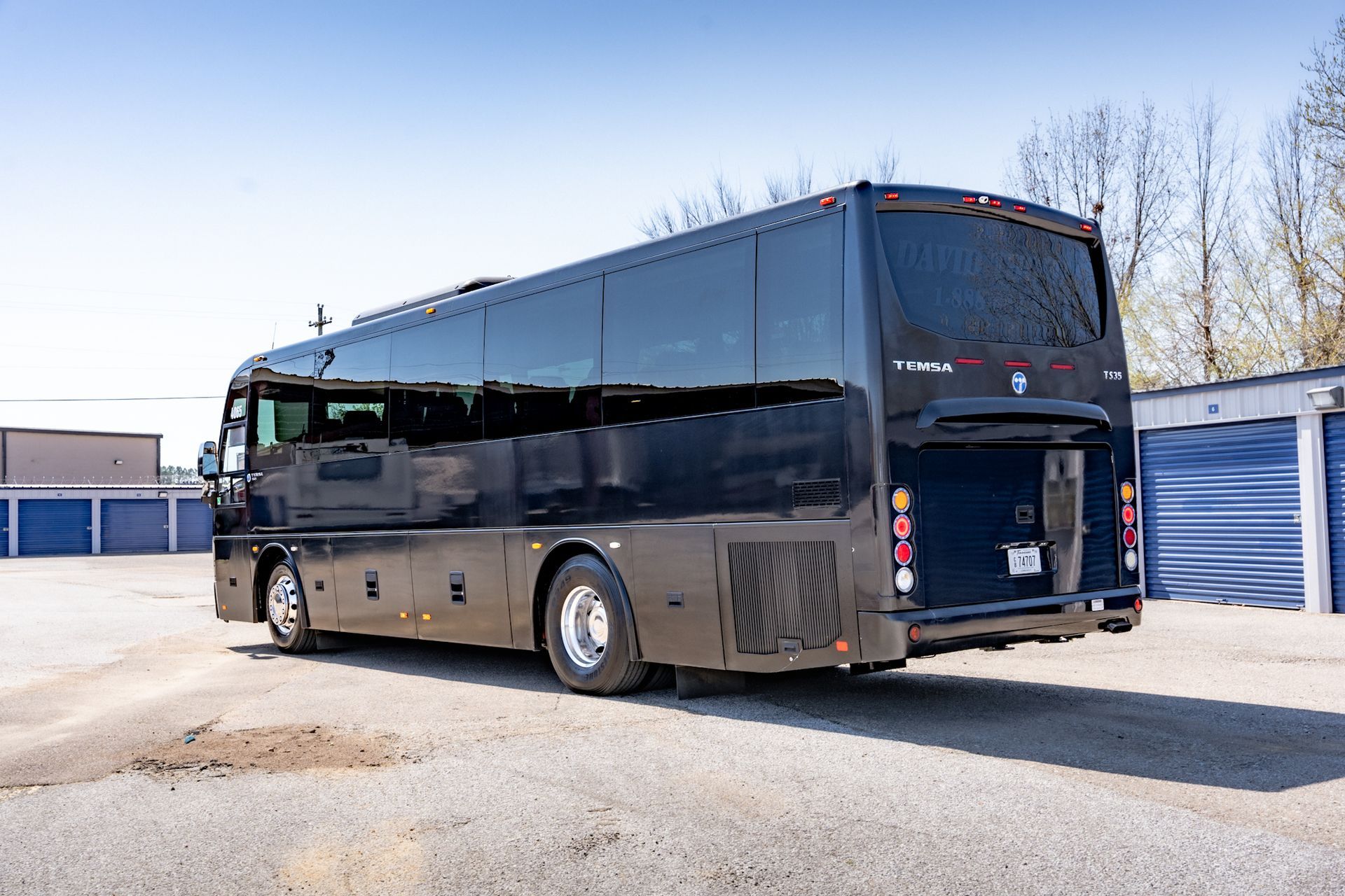 Black tour bus parked on gravel, rear door open, blue sky, storage units in background.