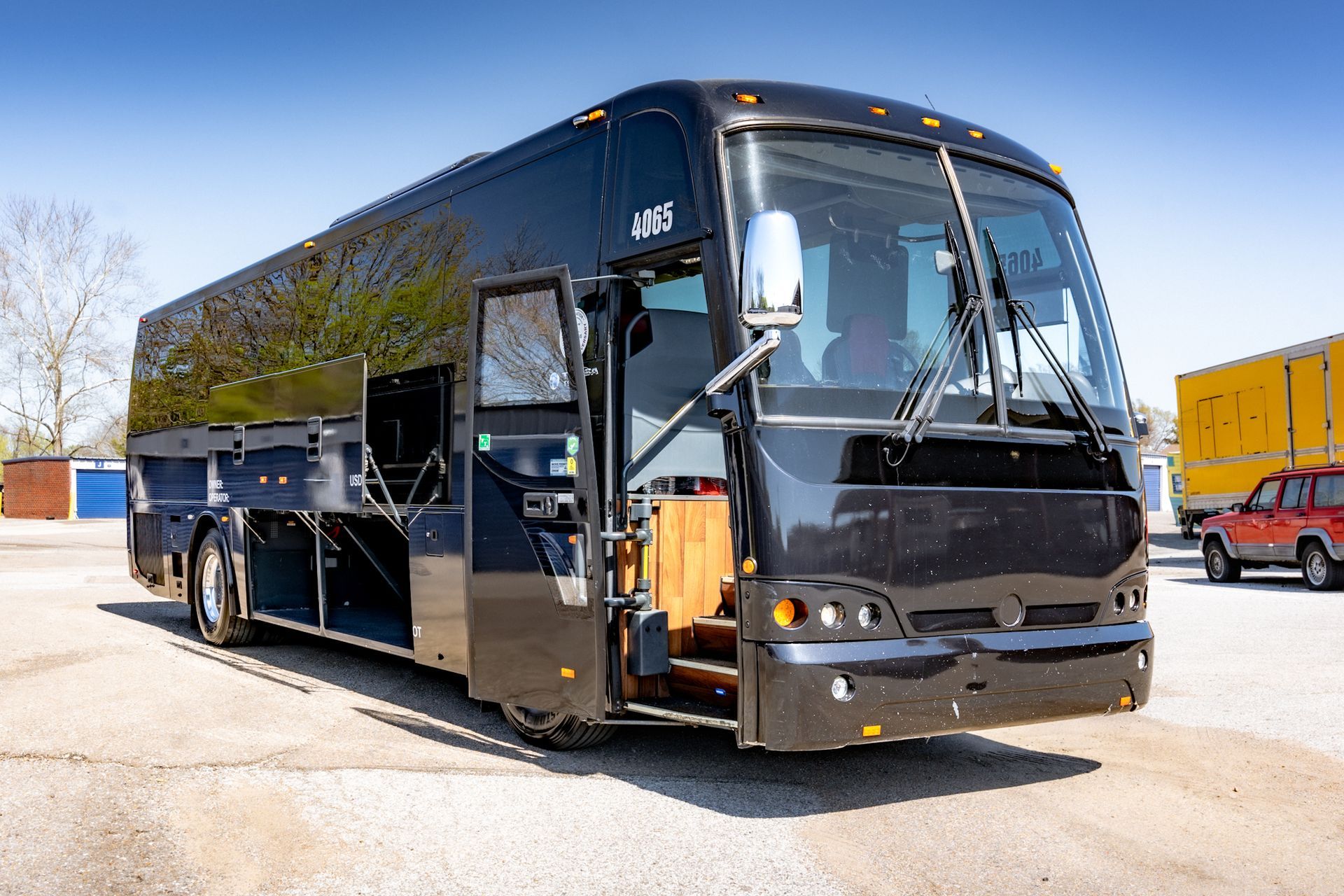 Black tour bus with open door, parked on a paved lot on a sunny day.
