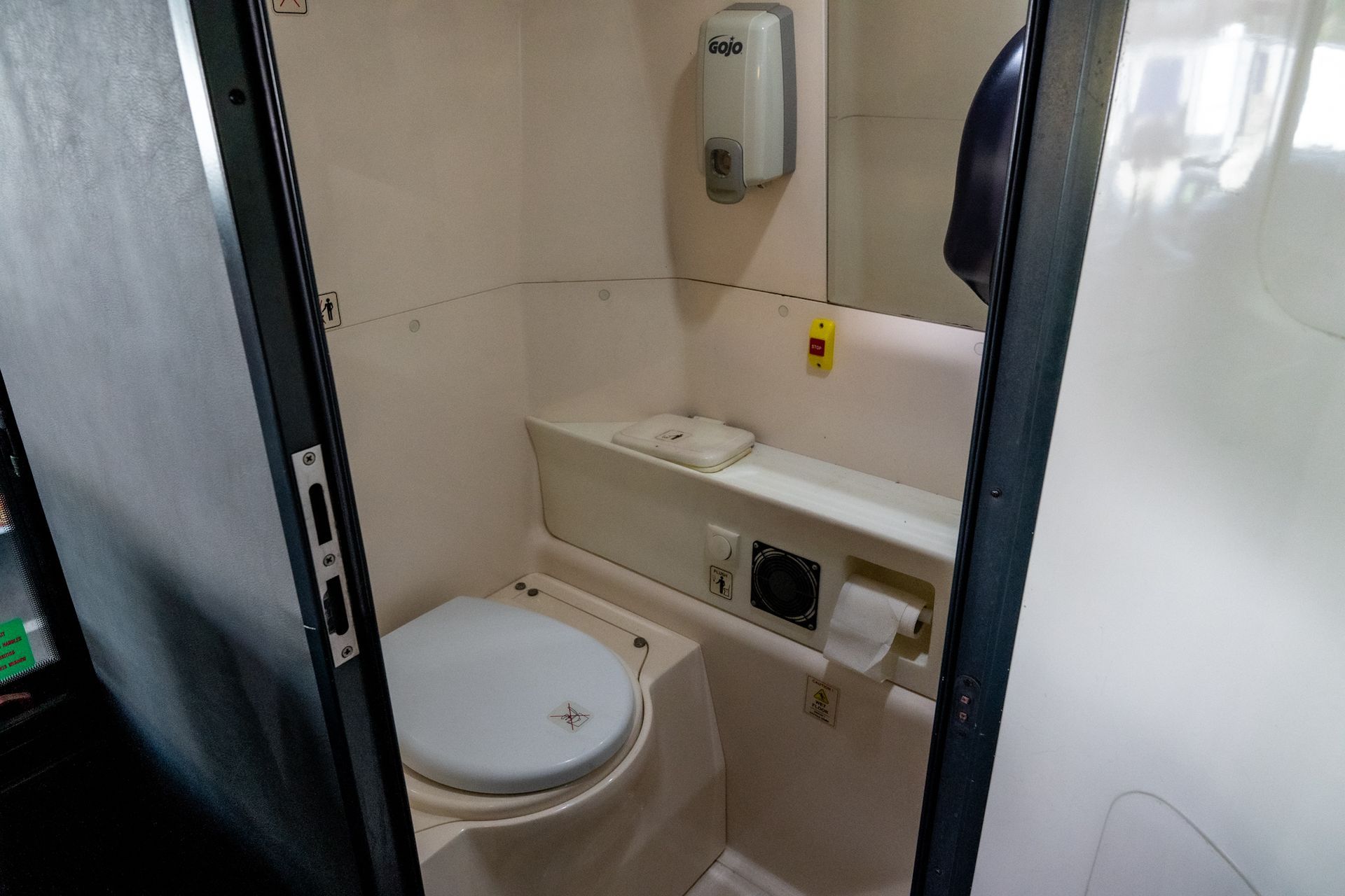 Small, enclosed bathroom in a vehicle, with a toilet, sink area, soap dispenser, and paper towel holder.