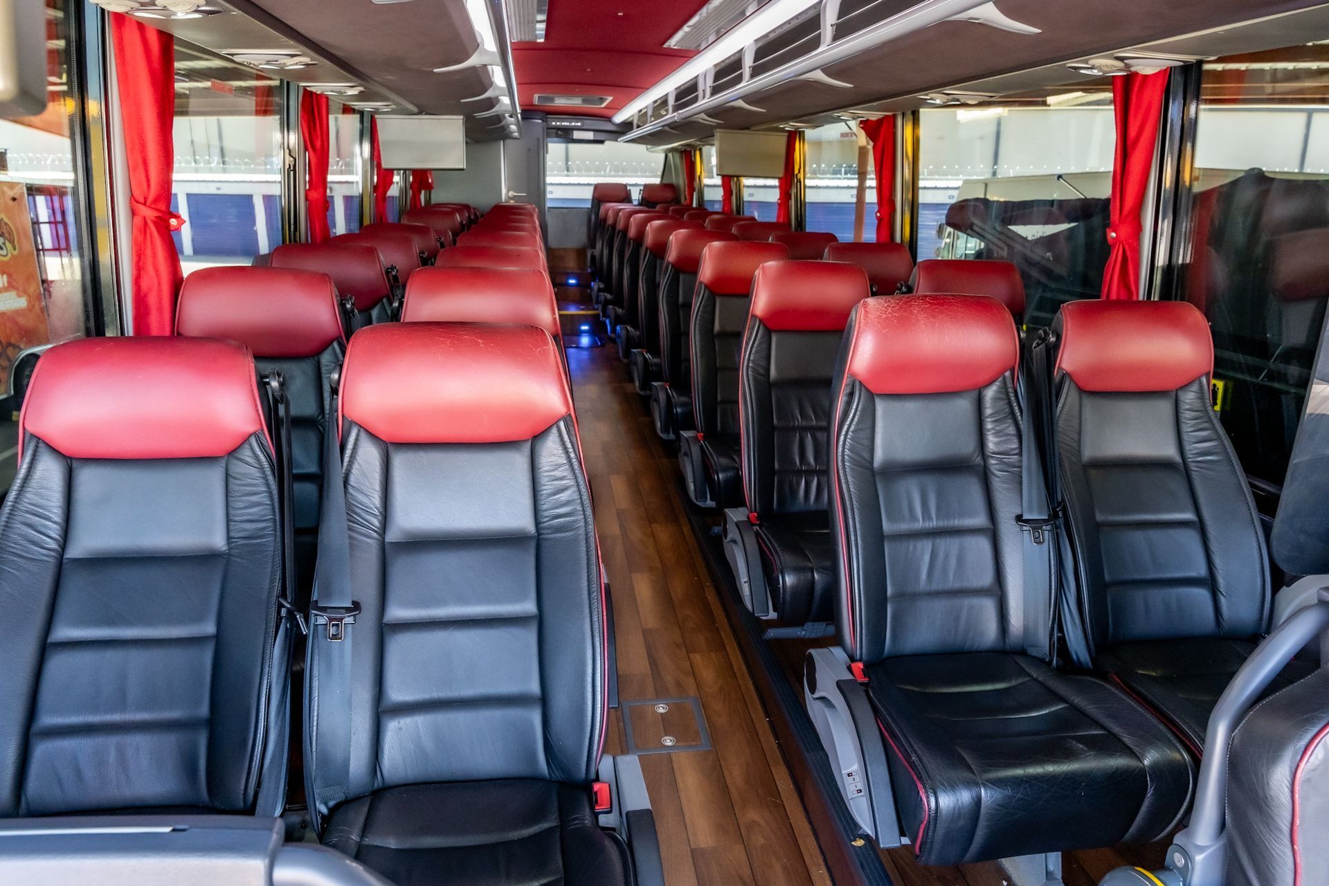 Interior view of a bus with rows of black and red seats and wooden flooring. Curtains hang on the windows.