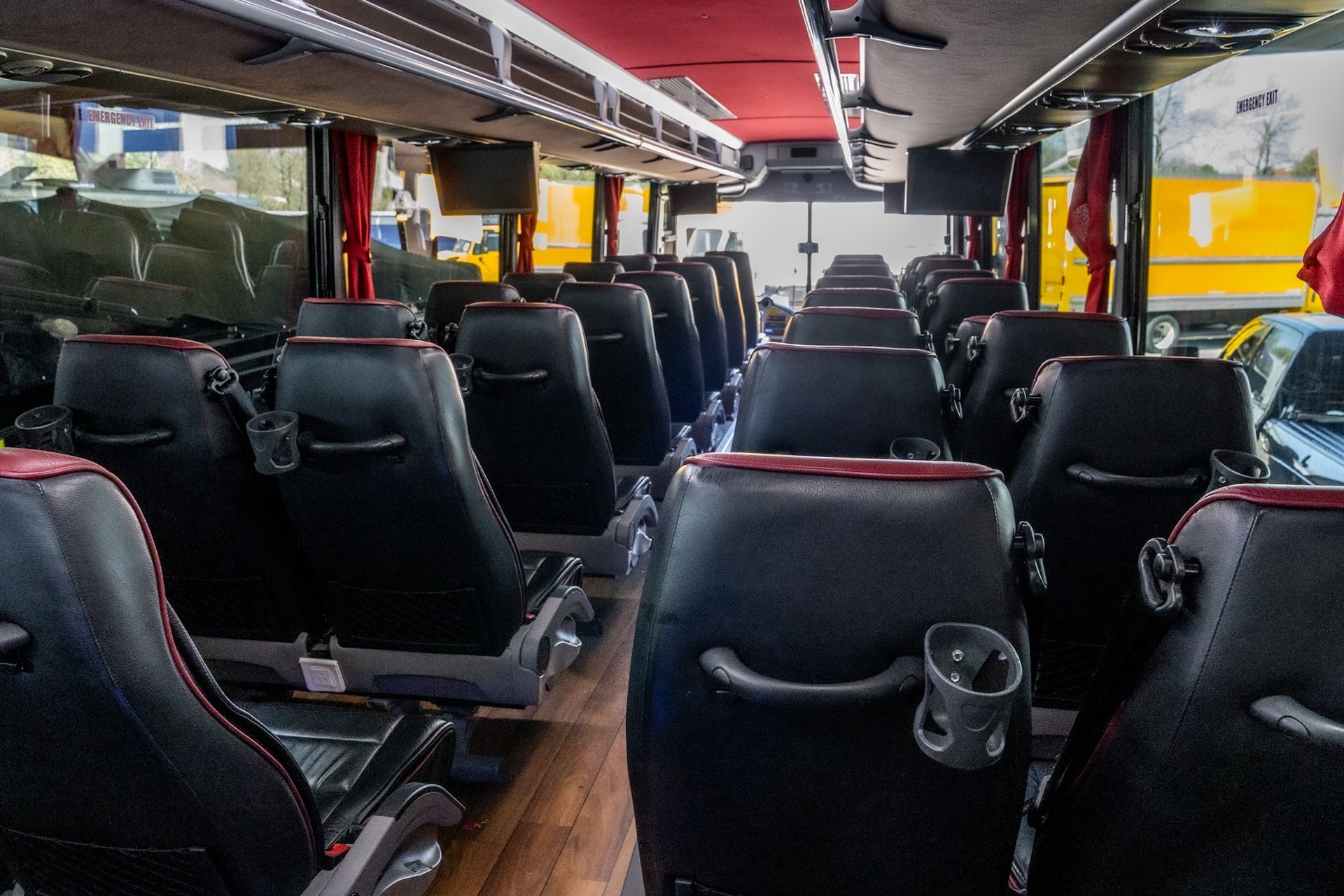 Interior of a coach bus with black seats, red accents, cup holders, and overhead screens.