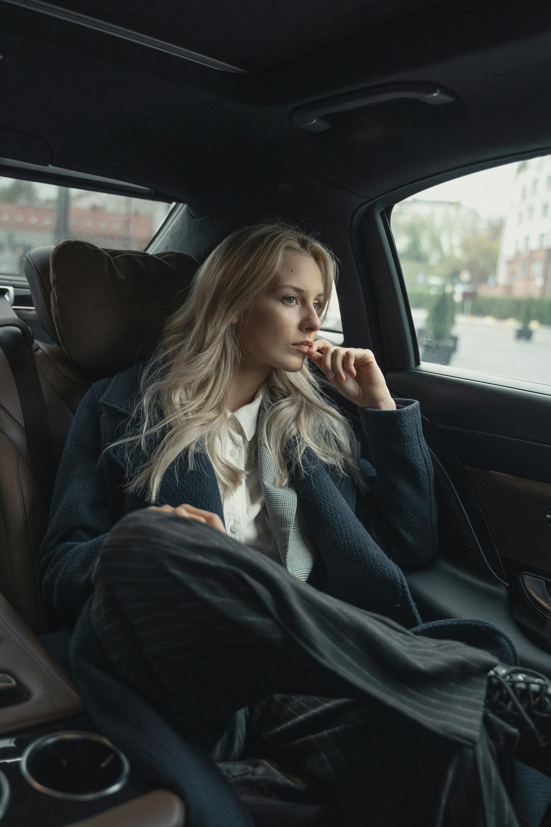 Woman in a car, looking out the window, wearing a coat and pants, thoughtful expression.