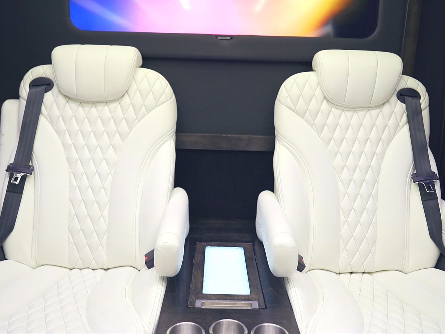 Two white quilted leather seats with seatbelts in a vehicle interior, centered on a console with cup holders.
