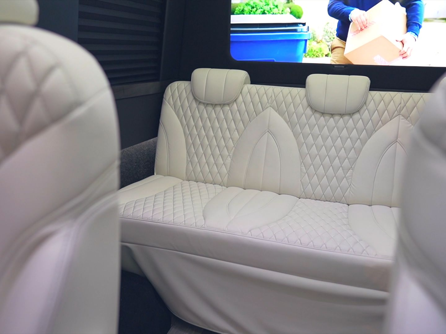 Interior van seats with white diamond-stitched upholstery, headrests, and a screen playing video.