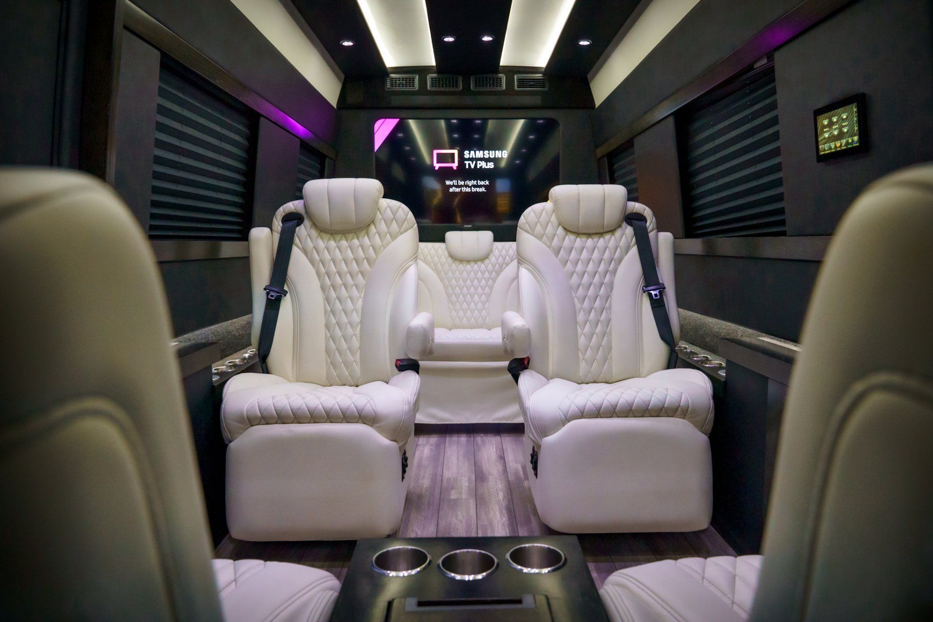 Interior of a luxury van with white leather seats, wood floor, and a large screen.
