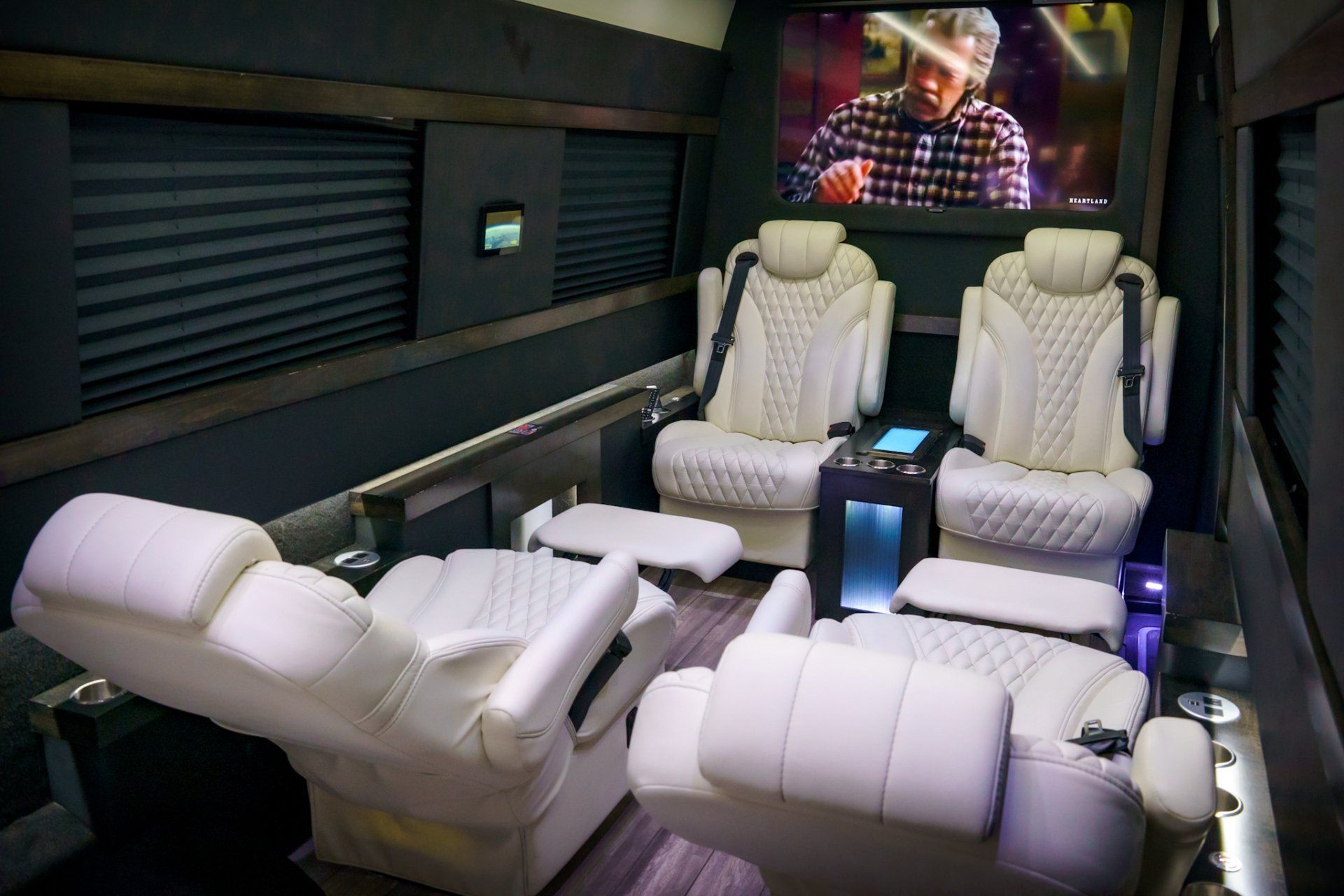 Interior of a luxury van with white quilted leather seats, TV screen, and dark gray walls.